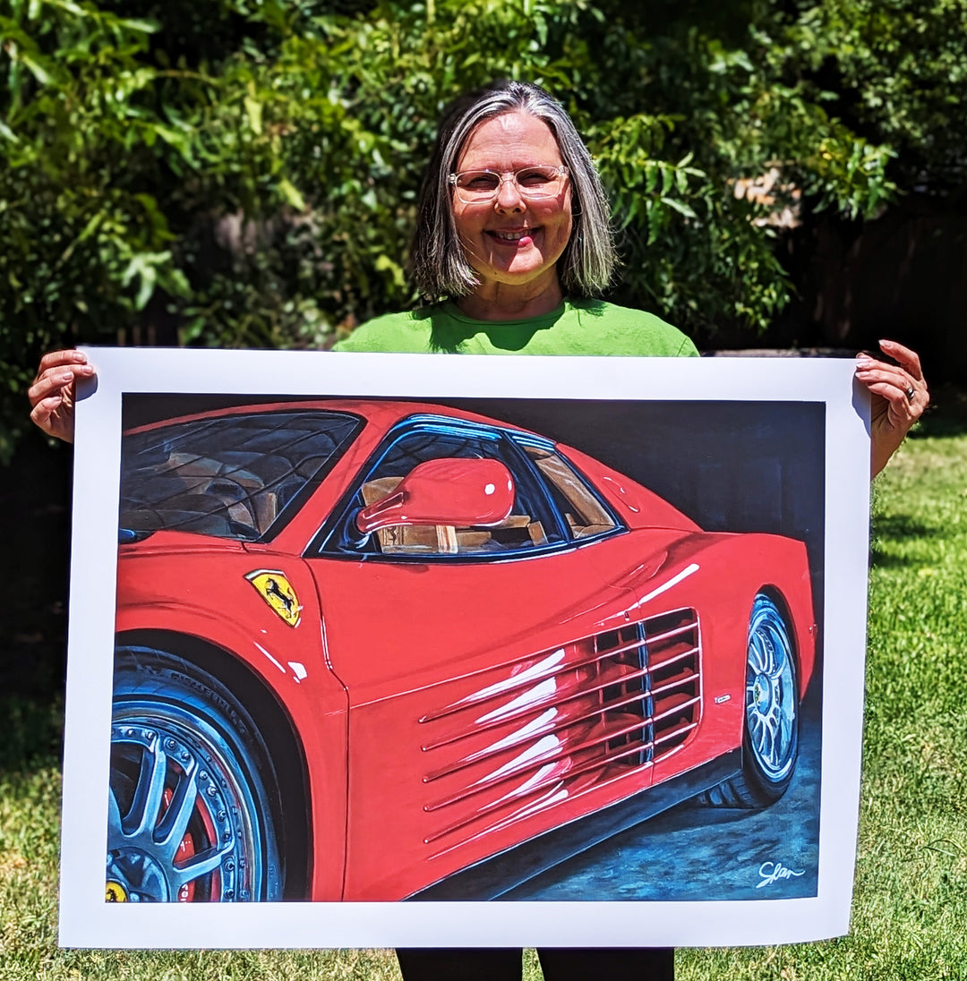 A woman holding up a painting of a red 1990 Ferrari Testarossa by Shan Fannin.