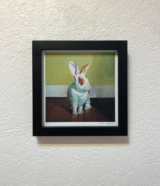 Benjamin Björklund | "Gwyneth" of a white rabbit with brown markings sitting on a dark surface against a green and yellow background. Created by Benjamin Björklund and printed with archival ink, this limited edition print is set in a black frame mounted on a textured white wall.