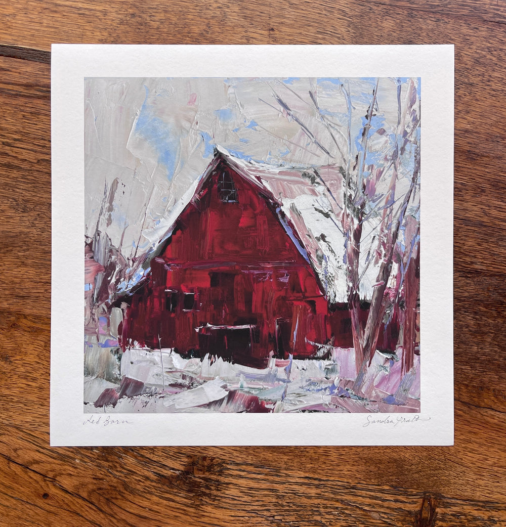 Sandra Pratt's "Winter Solstice" limited edition print by Lindsey Kustusch features a red barn in a snowy winter landscape with bare trees, displayed on a wooden surface.