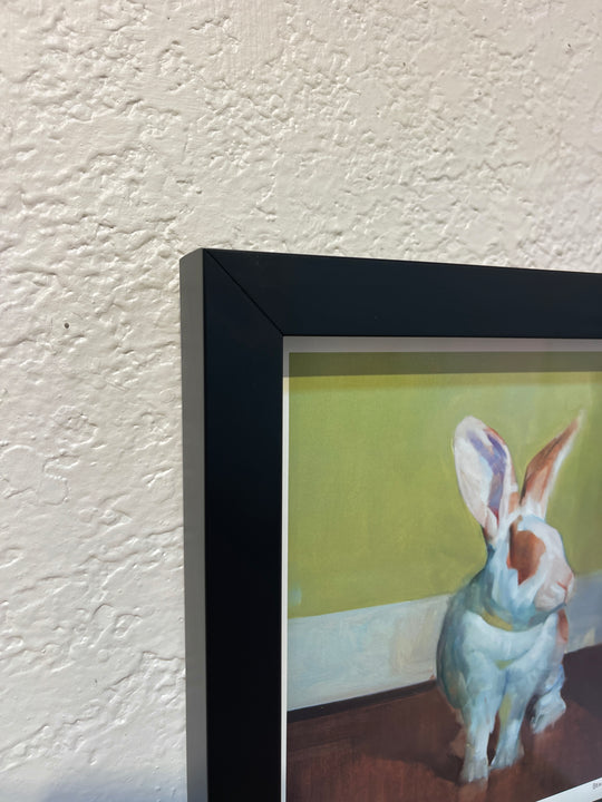 A close-up view of a framed limited edition print on a textured wall, showing the edge of the frame and a partial image of a rabbit with a green background. This piece by Benjamin Björklund is brought to life with rich archival ink. Product Name: Benjamin Björklund | "Gwyneth" Brand Name: Benjamin Björklund