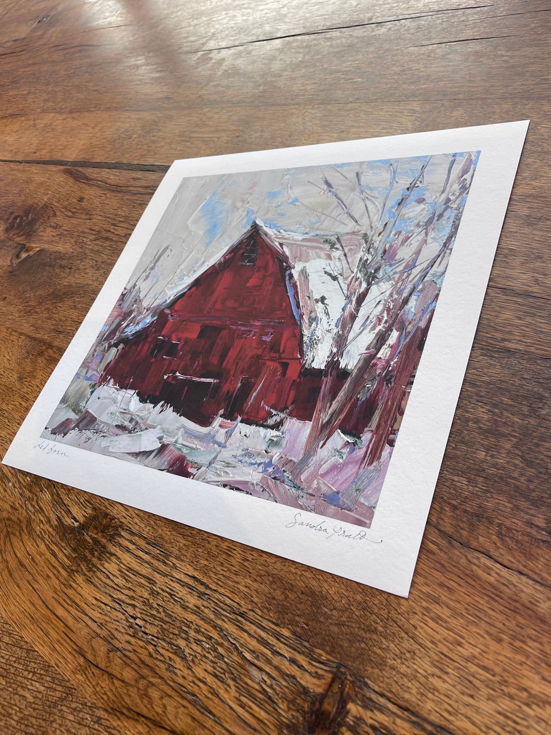 Sandra Pratt’s "Winter Solstice" limited edition print, offered by Lindsey Kustusch, features expressive brushstrokes of a snowy landscape with a red barn, set on a wooden surface and signed in the bottom right corner.