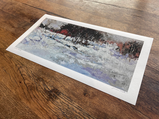 The Sandra Pratt | Winter Harmony limited edition print showcases a snowy landscape with a red building, printed on premium matte fine art paper and displayed on a wooden surface.